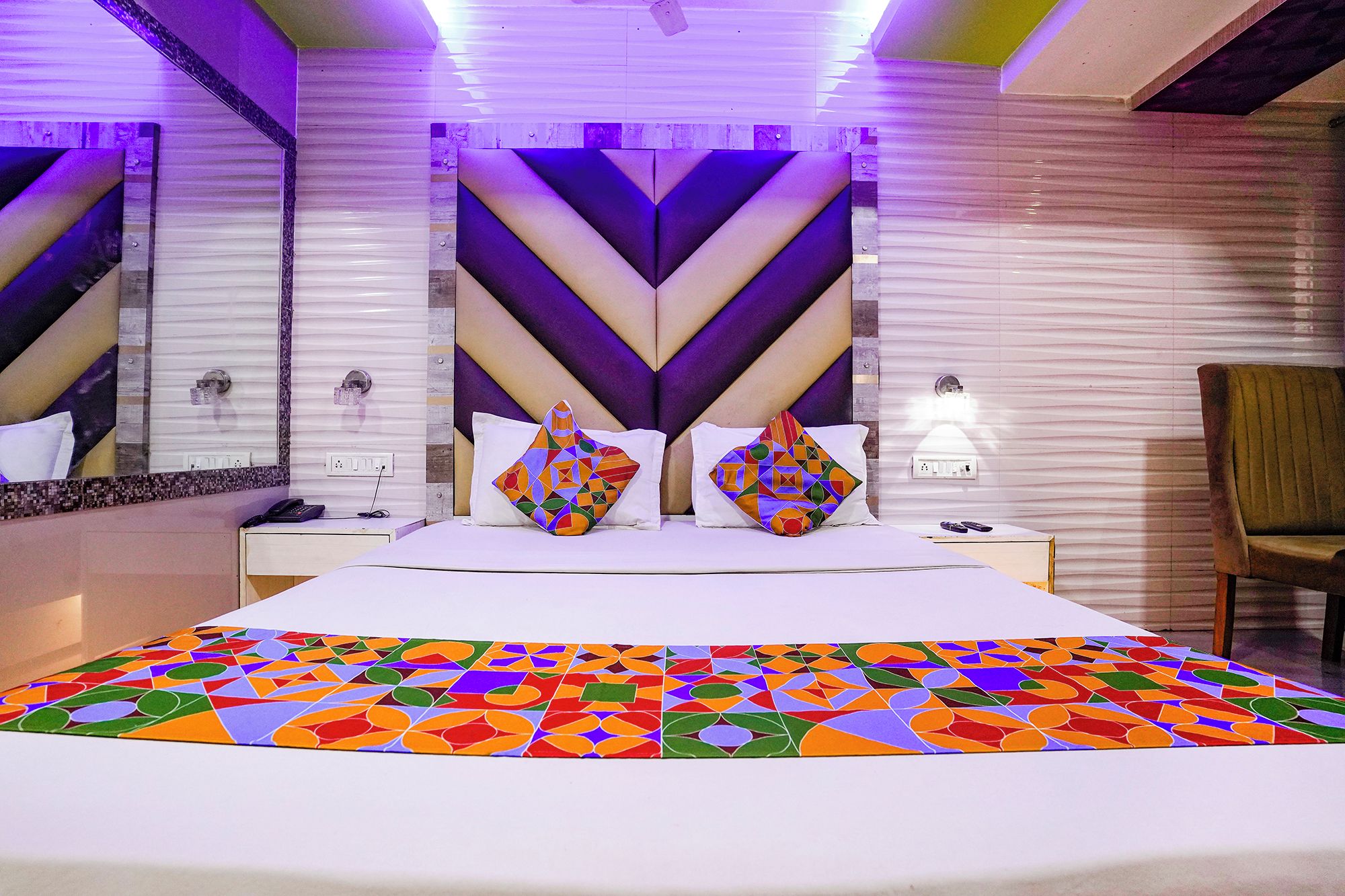 A modern hotel room with a large bed, two pillows, a nightstand, a chair, and a patterned rug.