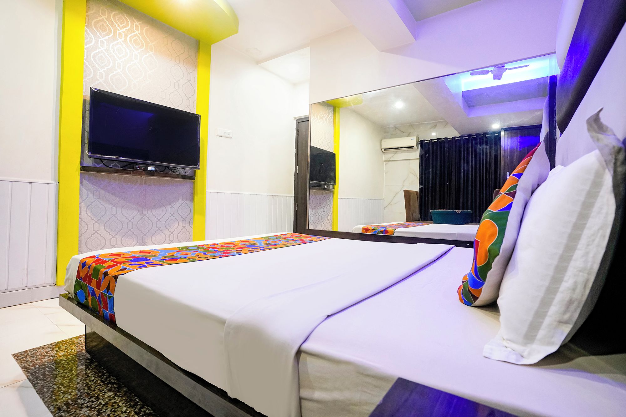 A hotel room with a bed, a flat-screen TV mounted on the wall, a mirror, and a ceiling fan.