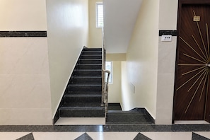 Una escalera con barandilla, una puerta con placa de número y un interruptor de luz en la pared.