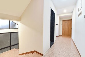 A hallway with a carpeted floor, a wooden door, and a window with blinds. - FabHotel The Samsara (Ujjain)