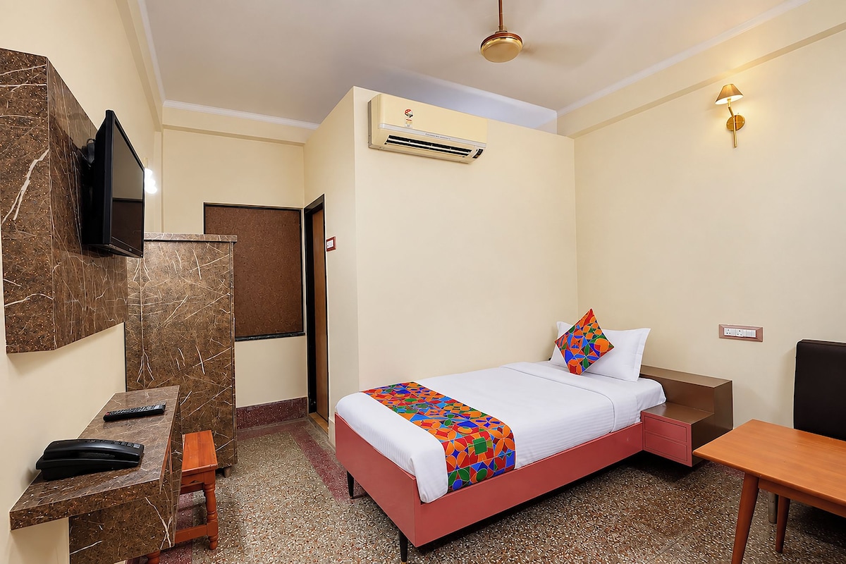 A hotel room with a bed, bedside table, desk, TV, air conditioning unit, and a wall-mounted light fixture.