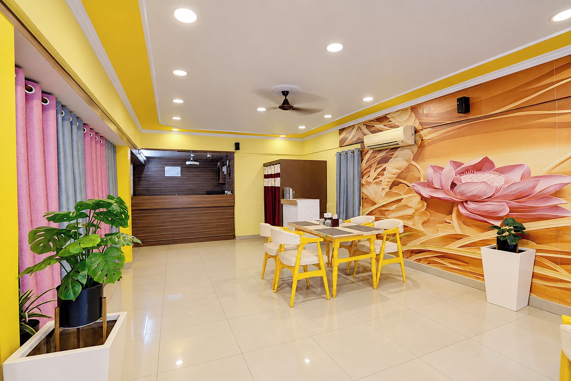 Un vestíbulo de hotel con mostrador de recepción, zona de comedor con sillas amarillas y un gran mural floral en la pared.