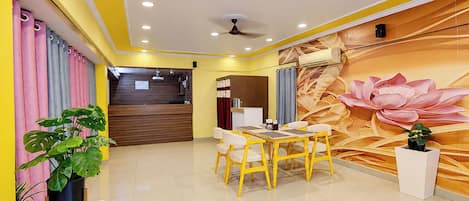 Un vestíbulo de hotel con mostrador de recepción, zona de comedor con sillas amarillas y un gran mural floral en la pared.