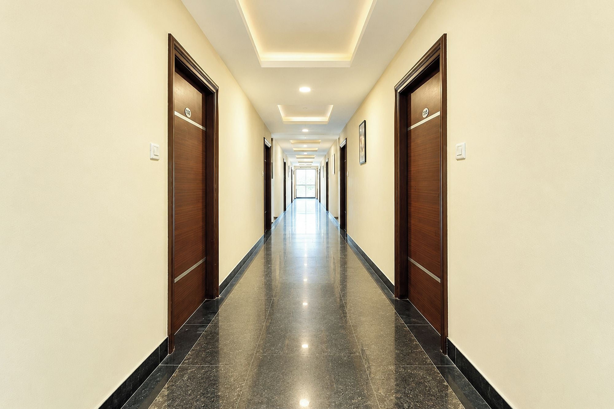 Un largo y estrecho pasillo de hotel con puertas a ambos lados, suelo de baldosas e iluminación empotrada en el techo.