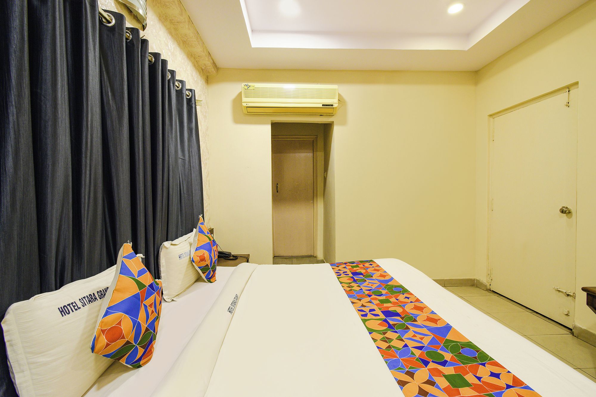 A hotel room with a bed, curtains, an air conditioning unit, a door, and a patterned runner.
