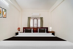A hotel room with a bed, a window with curtains, an air conditioning unit, and a nightstand with a lamp.