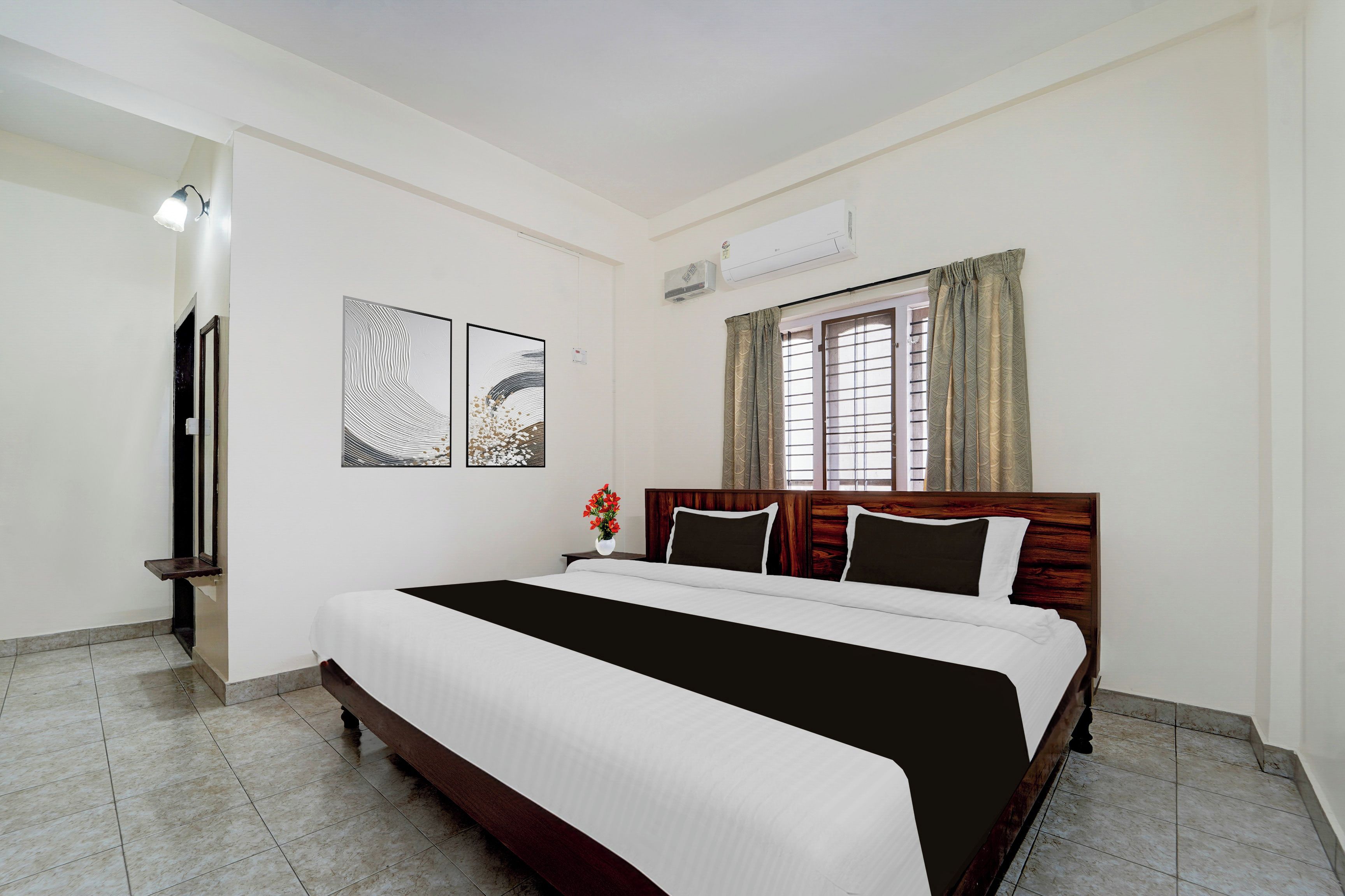 A hotel room with a large bed, a window with curtains, a wall-mounted air conditioner, and artwork on the wall.