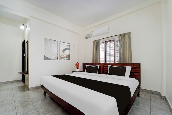 A hotel room with a large bed, a window with curtains, a wall-mounted air conditioner, and artwork on the wall.