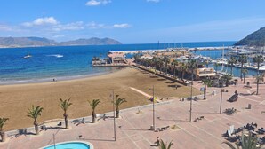 Blick auf einen Strand mit Swimmingpool, Palmen und einem Yachthafen mit Booten.