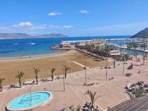 Vista sulla spiaggia con piscina, palme e un porto turistico con barche.