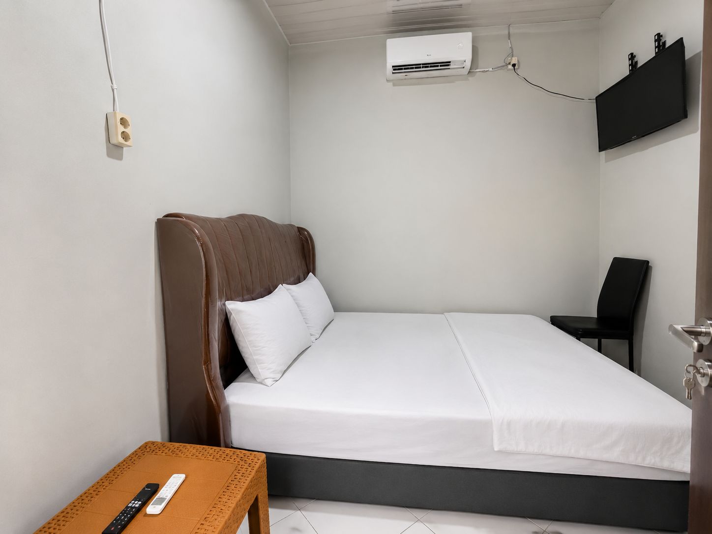 A single bed with a headboard, a bedside table with a remote control, a chair, and an air conditioning unit mounted on the wall.