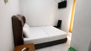 A small, single-bed room with a bed, a chair, a table with a remote control, a trash bin, and a wall-mounted air conditioning unit.