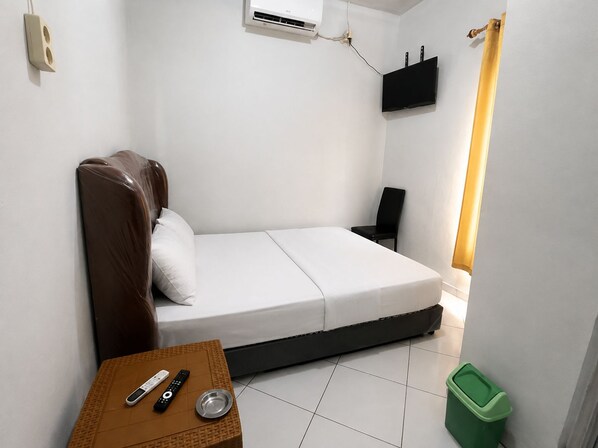A small, single-bed room with a bed, a chair, a table with a remote control, a trash bin, and a wall-mounted air conditioning unit.