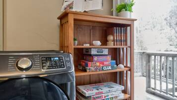 A room with a wooden bookshelf containing board games, a washing machine, and a window with a view of trees.
