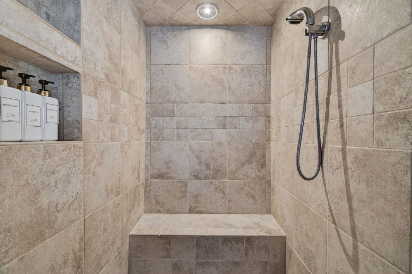 A shower area with beige tiles, a handheld showerhead, and a shelf with toiletries.