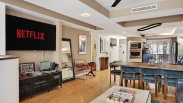 A modern living room with a large flat-screen TV, wooden flooring, a dining area with a wooden table and chairs, and a kitchen with built-in appliances.