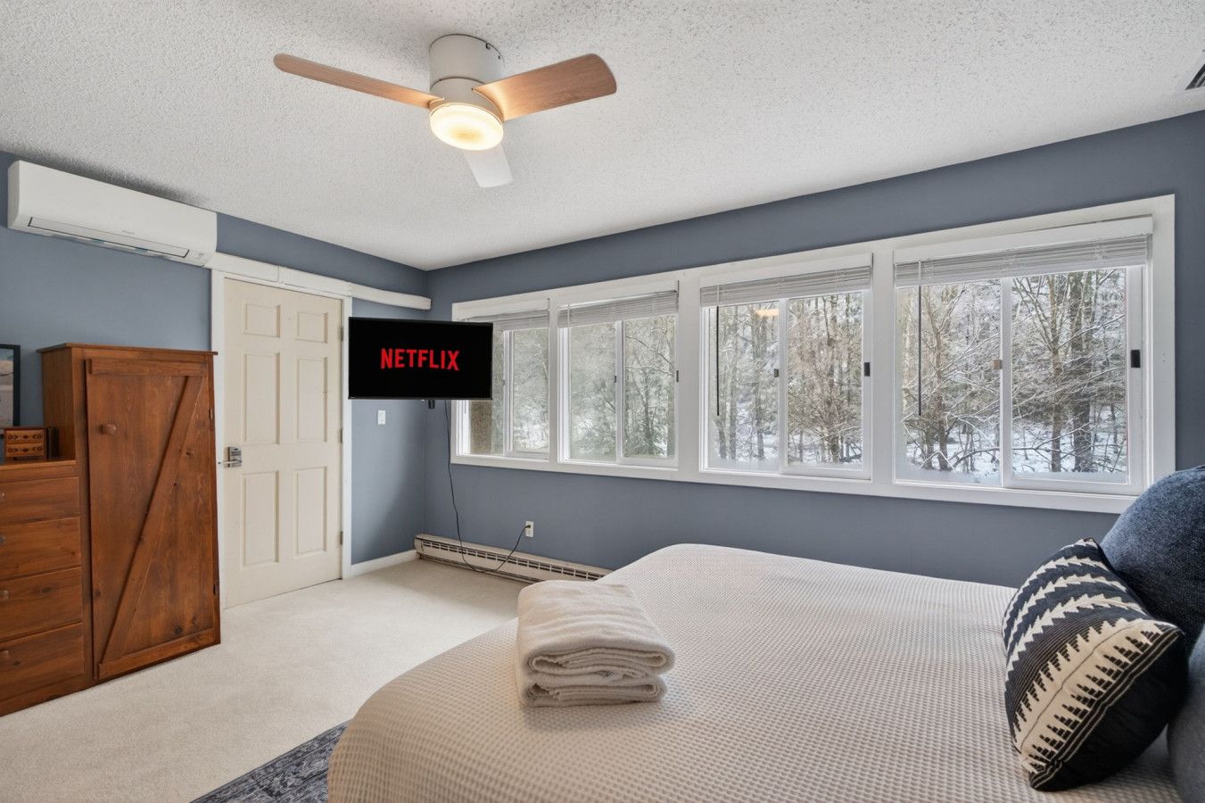 A bedroom with a bed, a ceiling fan, a TV mounted on the wall, and a window with a view of trees.