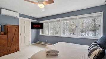 A bedroom with a bed, a ceiling fan, a TV mounted on the wall, and a window with a view of trees.