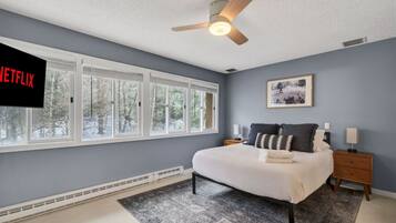 A bedroom with a bed, nightstands, a ceiling fan, a window with blinds, and a wall-mounted TV.