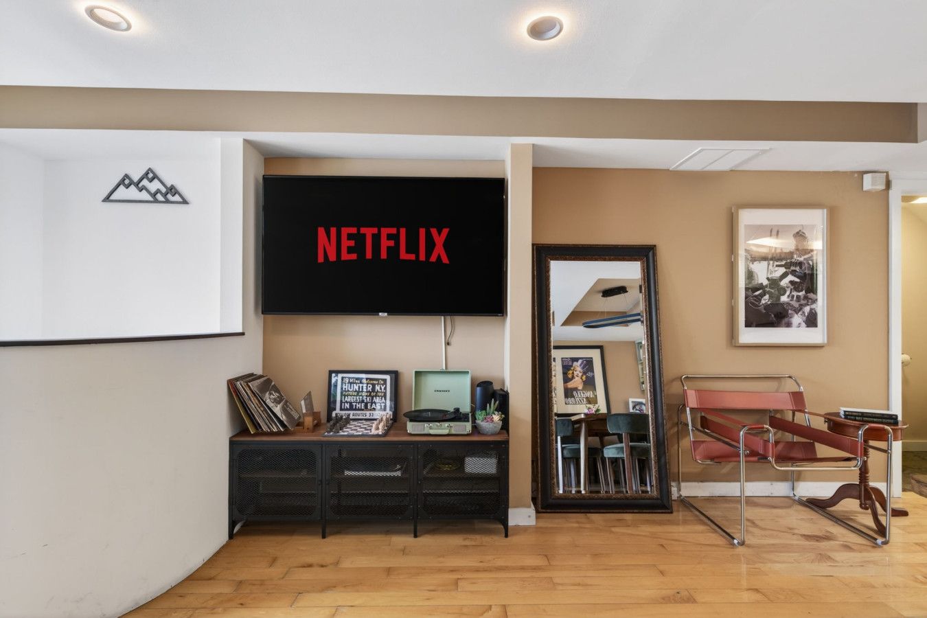 Une pièce avec un téléviseur fixé au mur qui affiche le logo « NETFLIX », une bibliothèque contenant divers objets, un miroir et un fauteuil rouge.