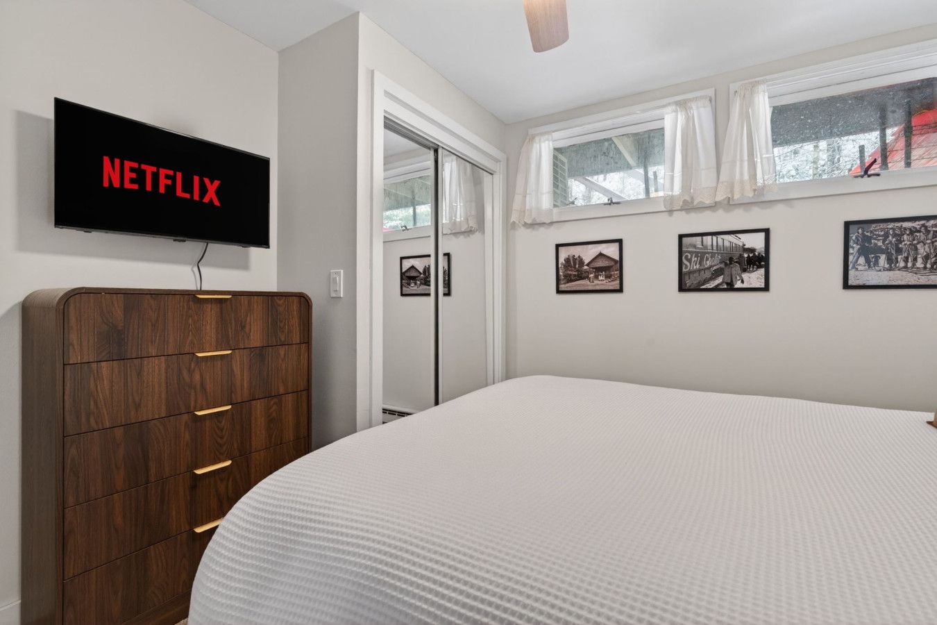 Une chambre avec un grand lit, une commode en bois, un téléviseur fixé au mur qui affiche « NETFLIX », et trois tableaux encadrés accrochés au mur.