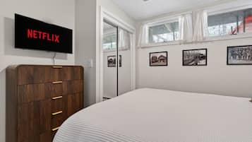 Une chambre avec un grand lit, une commode en bois, un téléviseur fixé au mur qui affiche « NETFLIX », et trois tableaux encadrés accrochés au mur.