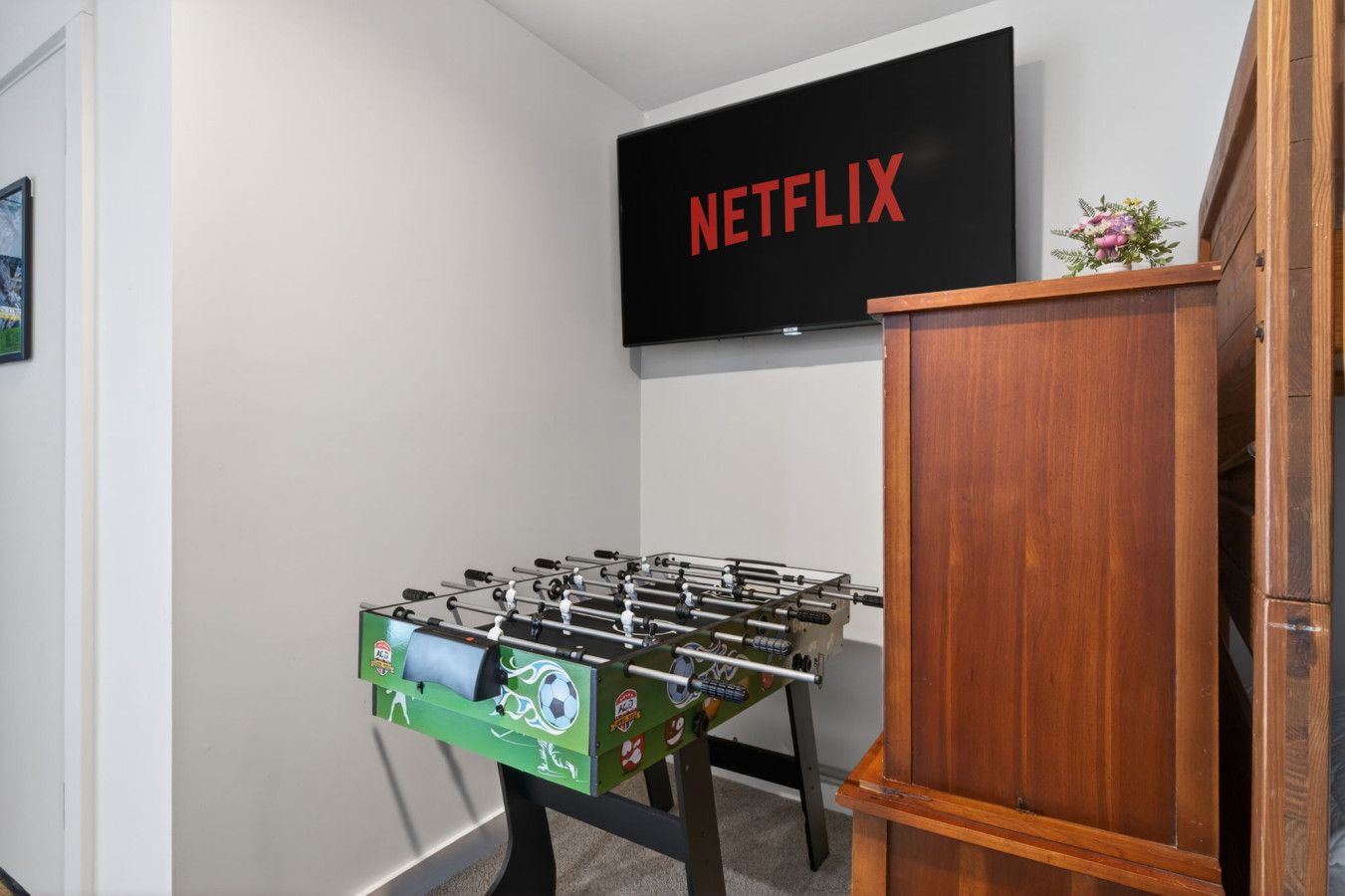 Une pièce comprenant un baby-foot, un meuble en bois et un téléviseur fixé au mur, sur lequel apparaît le logo de Netflix.