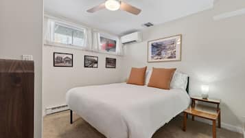 A bedroom with a bed, a bedside table with a lamp, a ceiling fan, a window with curtains, and framed pictures on the wall.