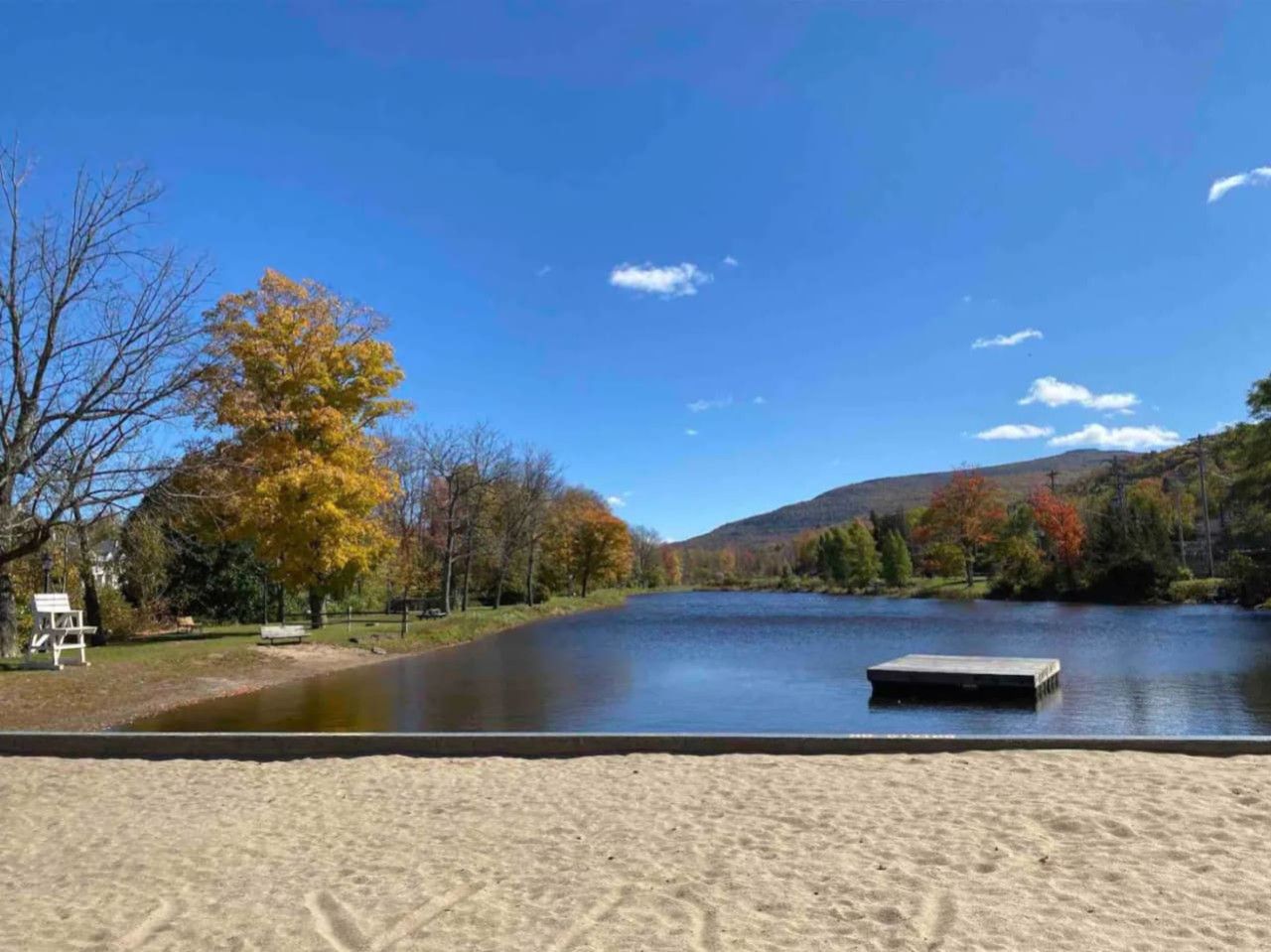 Un lac avec un quai, entouré d’arbres aux feuilles d’automne, sous un ciel bleu dégagé.