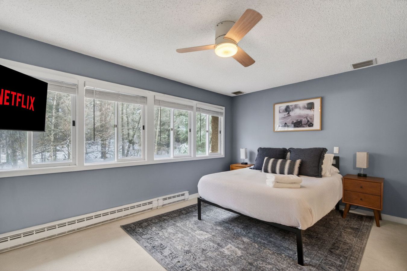 A bedroom with a bed, nightstands, a ceiling fan, a window with blinds, and a wall-mounted TV.