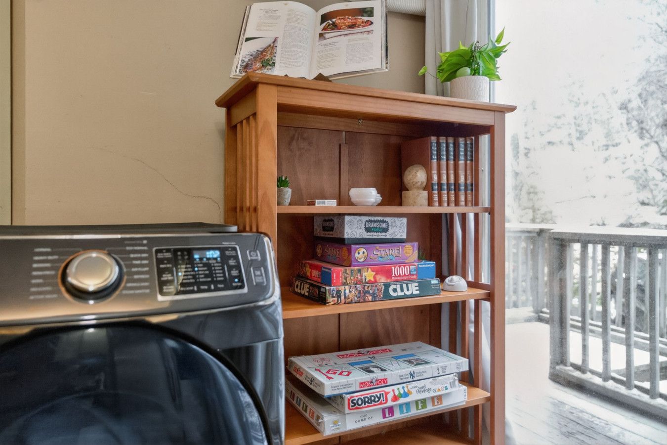 A room with a wooden bookshelf containing board games, a washing machine, and a window with a view of trees.