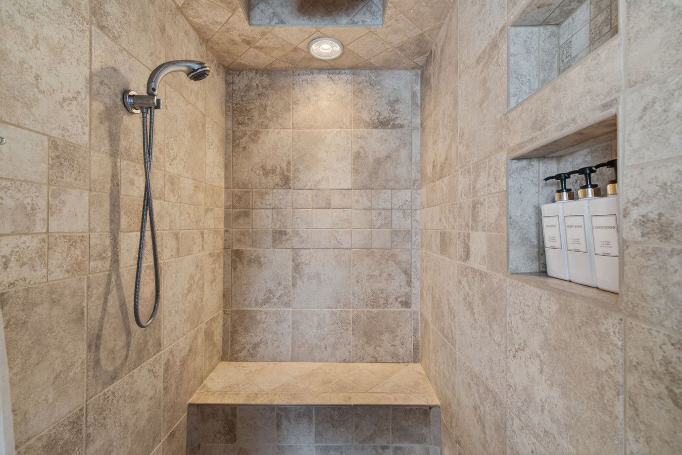 Une douche avec un pommeau de douche à main, une étagère intégrée pour les articles de toilette et un spot encastré au plafond.
