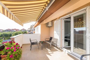 Ein Balkon mit gestreiftem Sonnensegel, einem Tisch, zwei Stühlen und einem Blumentopf mit rosa Blüten.