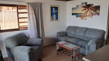Una sala de estar con un sofá gris, un sillón a juego, una mesa de centro de madera con mantel estampado y una pared decorada con marcos.