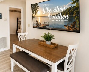 A dining area with a wooden table, white chairs, and a television displaying a lake vacation scene.
