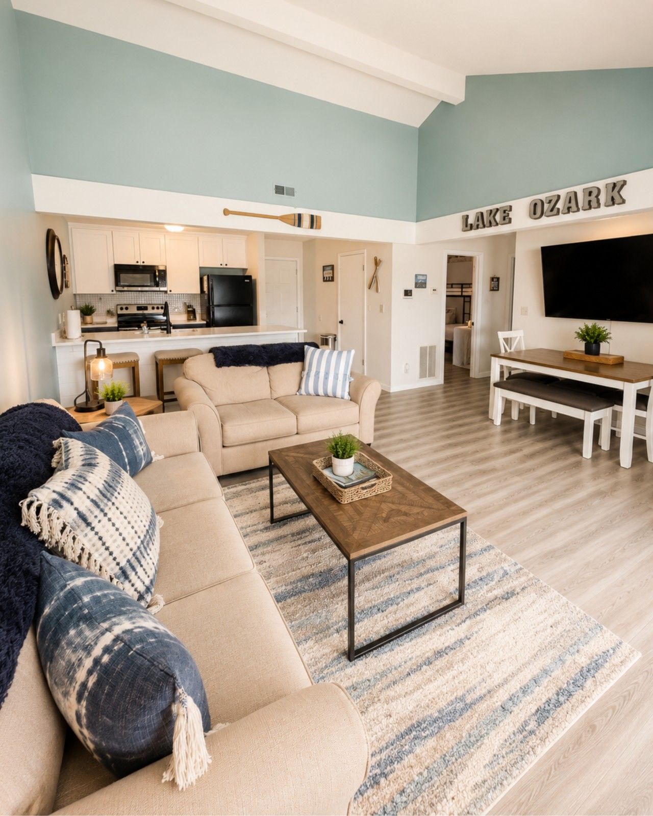 A living room with a sofa, coffee table, and a TV. There is a kitchen area with a microwave and stove. The room has a large window and a sign that reads 'LAKE OZARK'.