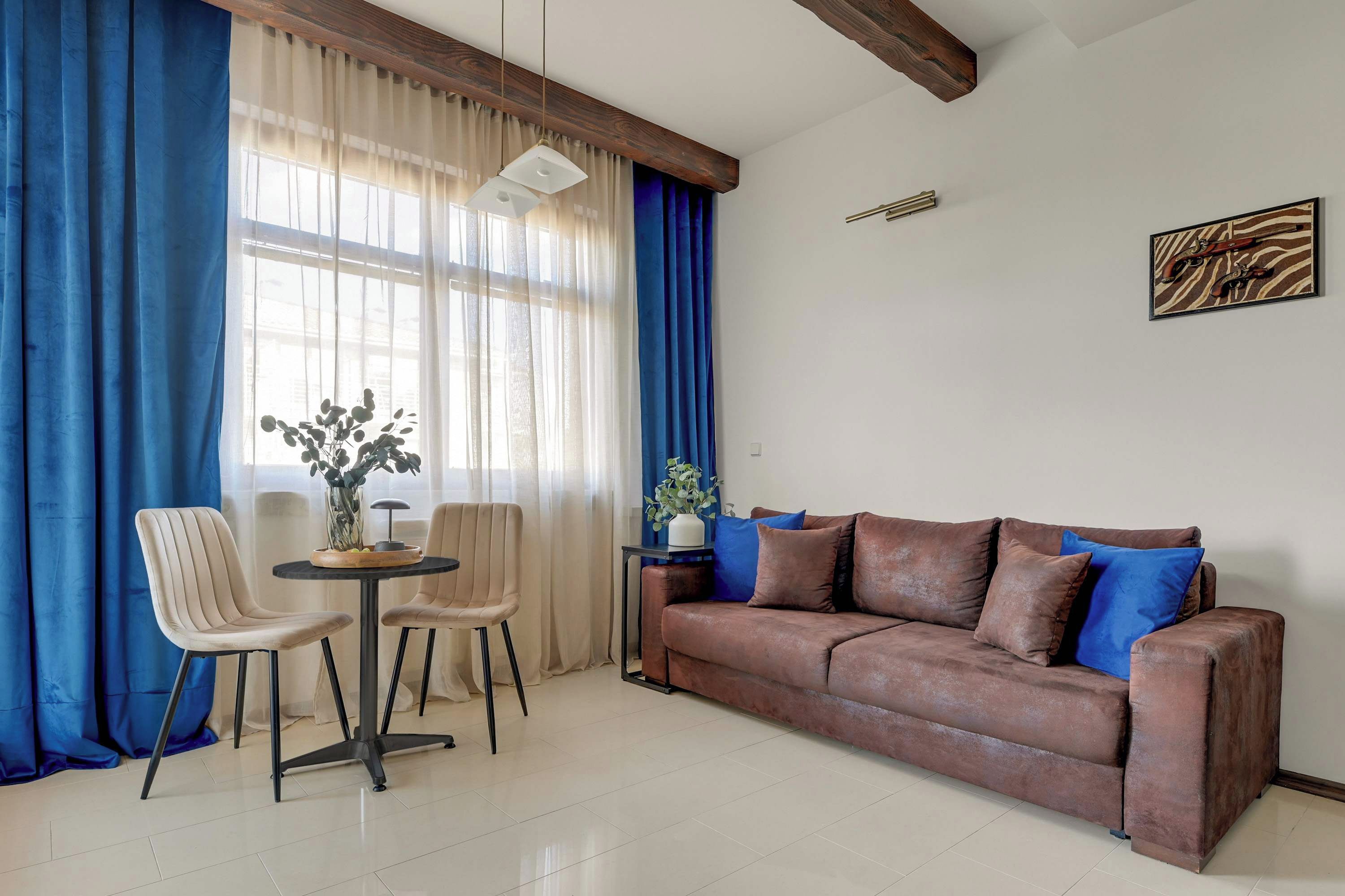 A living room with a brown sofa, two chairs, a small table, and a large window with blue curtains.