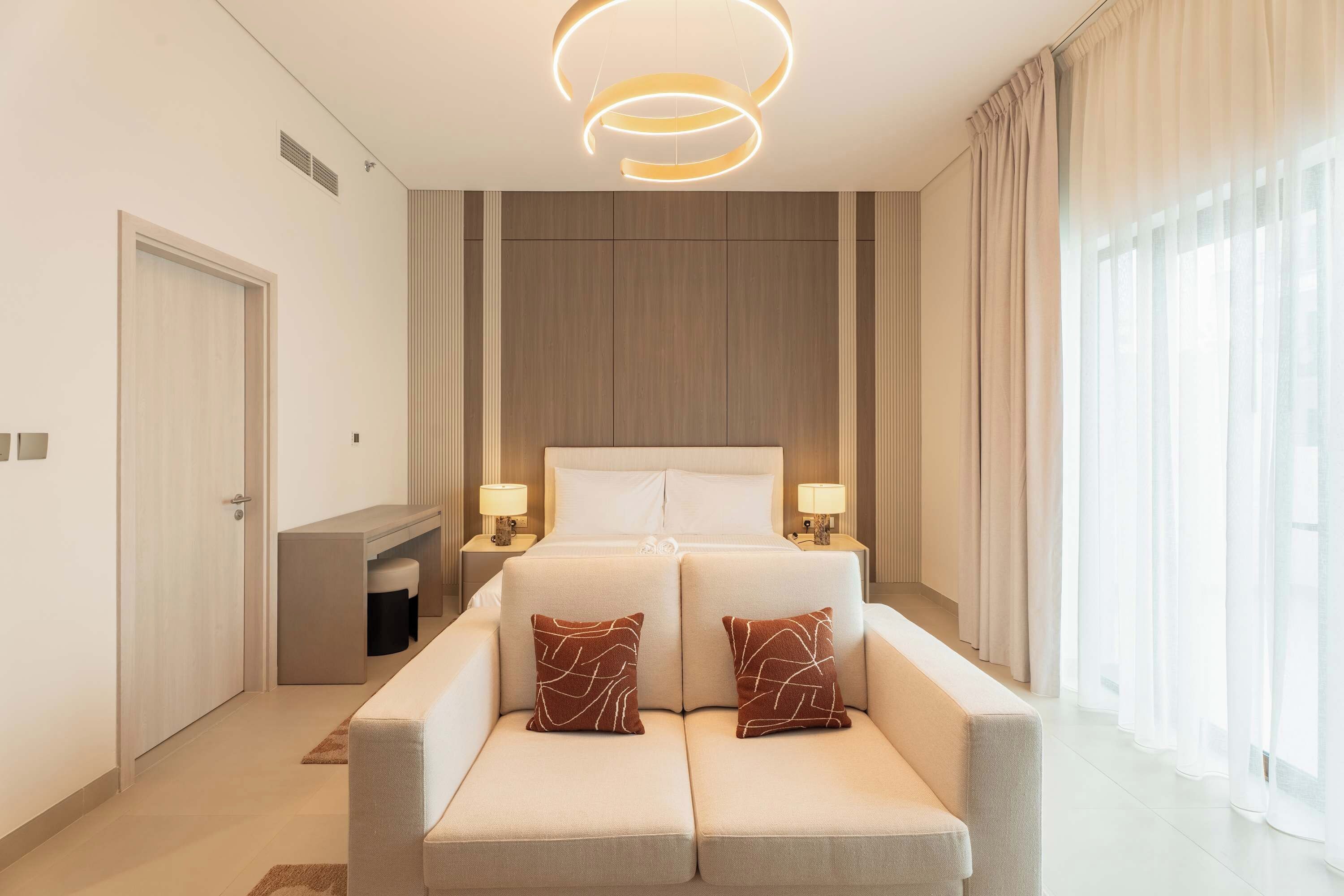 A modern hotel room with a large bed, two bedside tables with lamps, a sofa, and a large window with curtains.