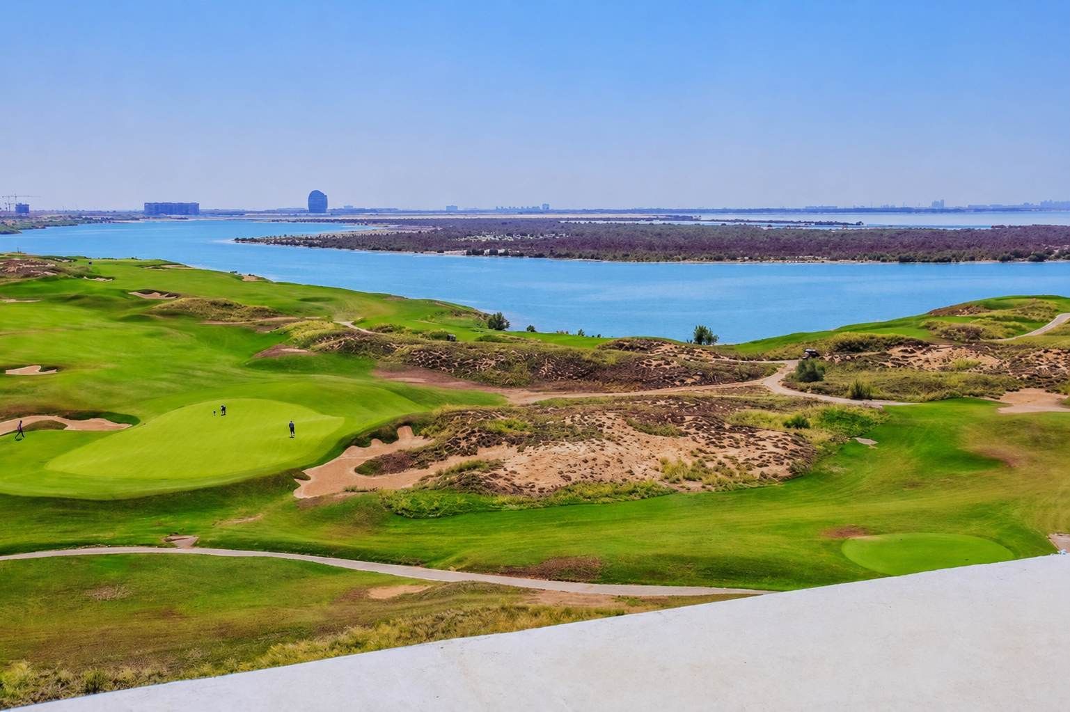 A golf course with bunkers, a water body, and distant buildings.