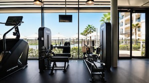 A gym with cardio equipment, weight machines, and a view of a pool and palm trees through large windows. - Vanilla Homes Modern Studio Yas Island (Abu Dhabi)