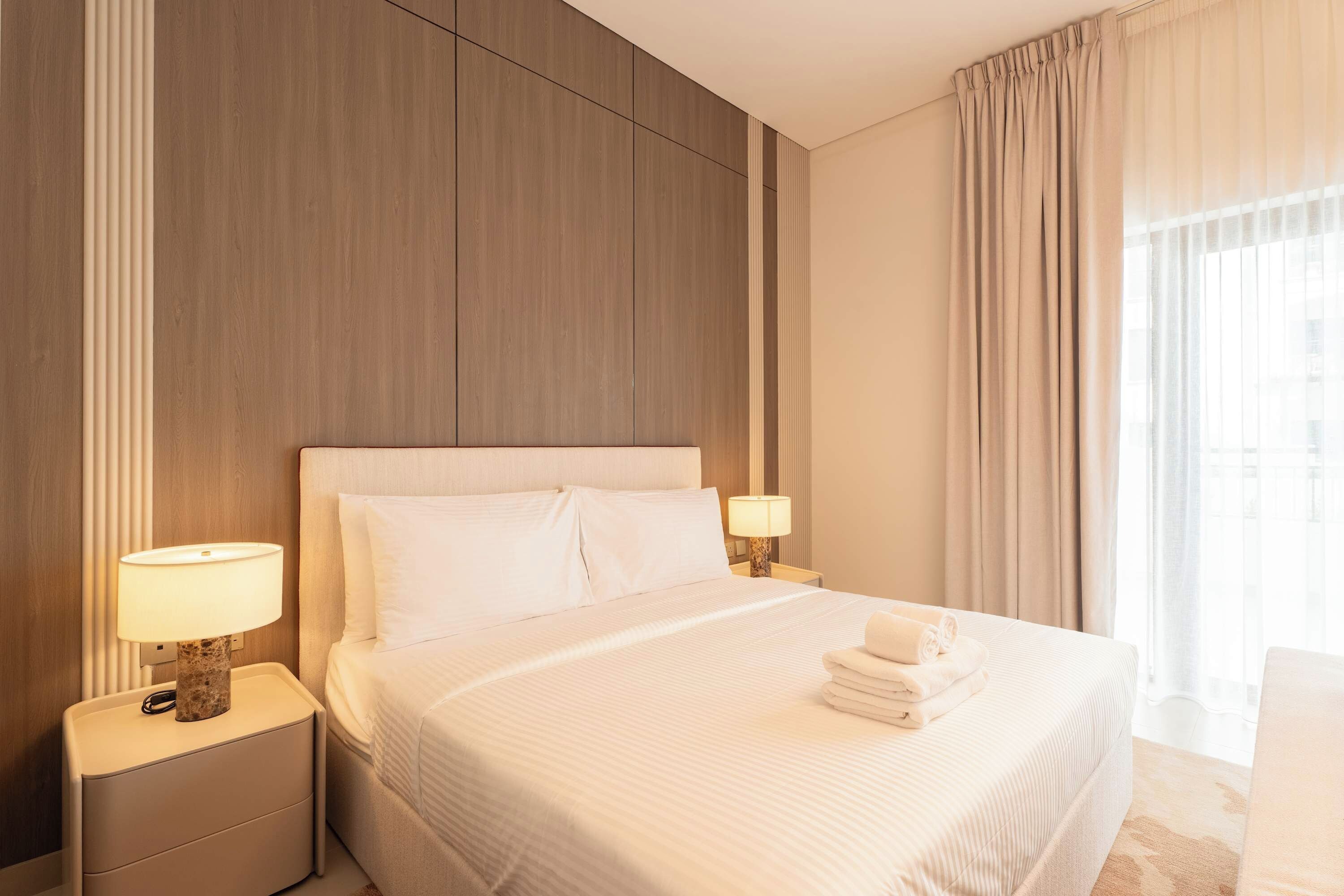 A hotel room with a large bed, two bedside tables with lamps, a headboard, and a view of the outdoors through the window.