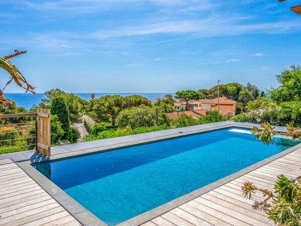 A rectangular swimming pool with a wooden deck surrounding it, set against a backdrop of a clear sky and a view of the sea and greenery. - Villa in Les Issambres With Swimming Pool (Roquebrune-sur-Argens)