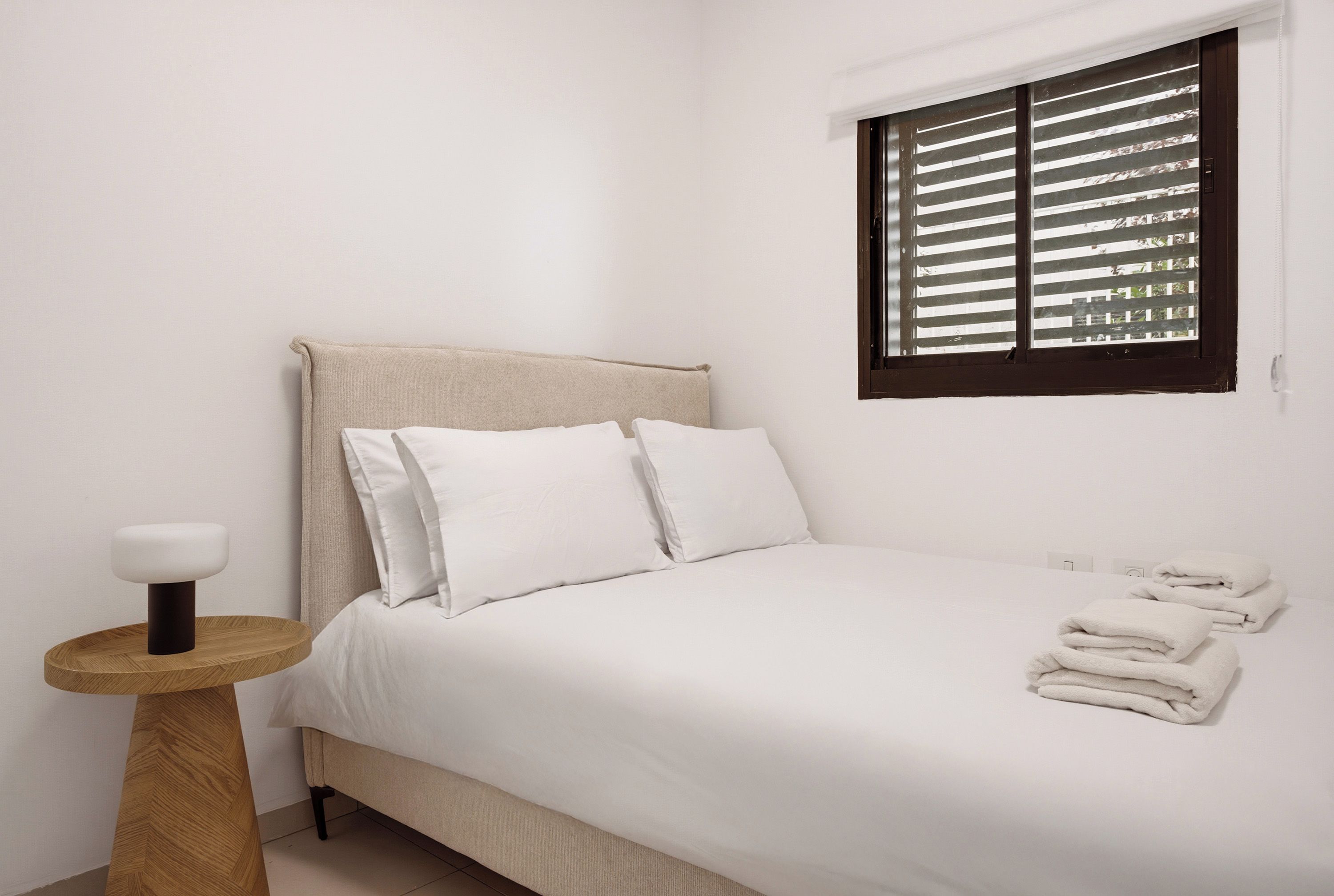 Quarto com cama, almofadas, mesa de cabeceira, janela com persianas e uma pilha de toalhas.