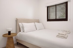 A bedroom with a bed, pillows, a bedside table, a window with blinds, and a towel stack. - Spacious 4BR Luxury l Balcony & Mamad (Petah Tikva)