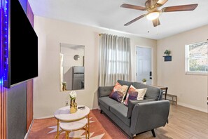 A living room with a grey sofa, a small round table, a ceiling fan, and a wall-mounted TV. - Your Ybor Base - WFH Pet Friendly - Full Kitchen (Tampa)