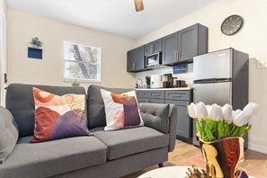 A living room with a grey sofa and decorative pillows, a kitchen with grey cabinets and a refrigerator, and a clock on the wall. - Your Ybor Base - WFH Pet Friendly - Full Kitchen (Tampa)