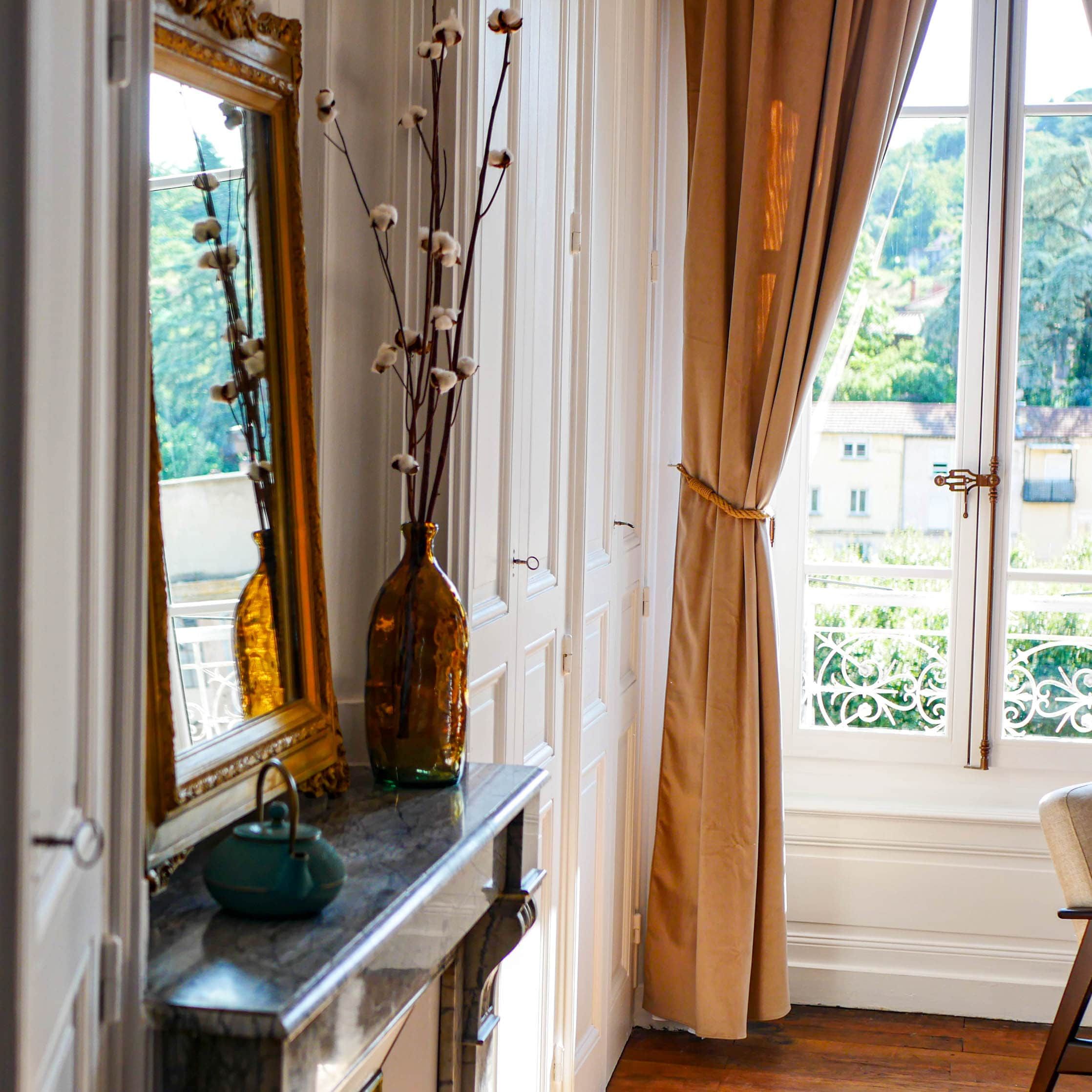 A room with a window, a mirror, a vase with branches, and a curtain.