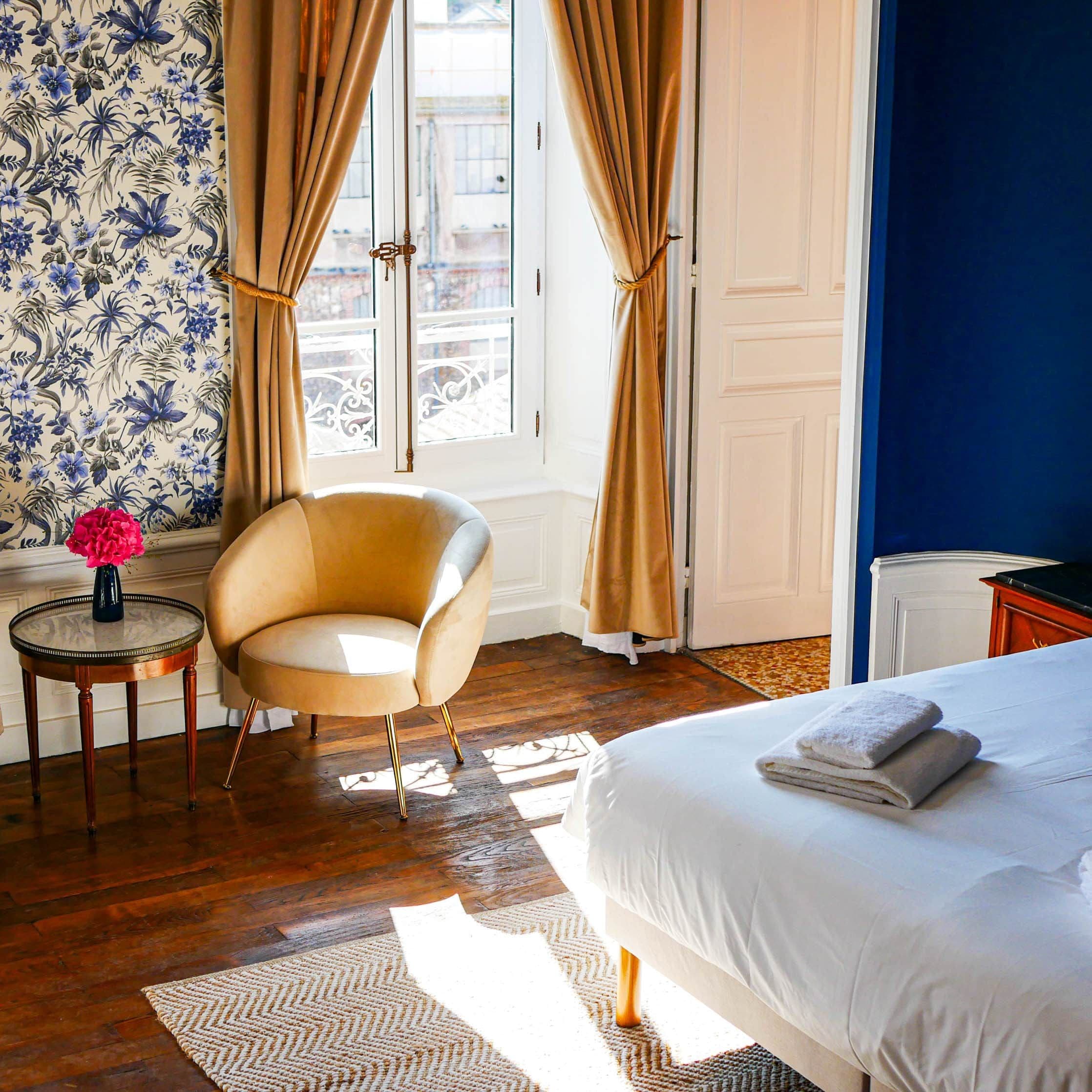Une chambre d’hôtel avec un lit, une chaise, une table ronde ornée d’un vase et une fenêtre avec des rideaux.