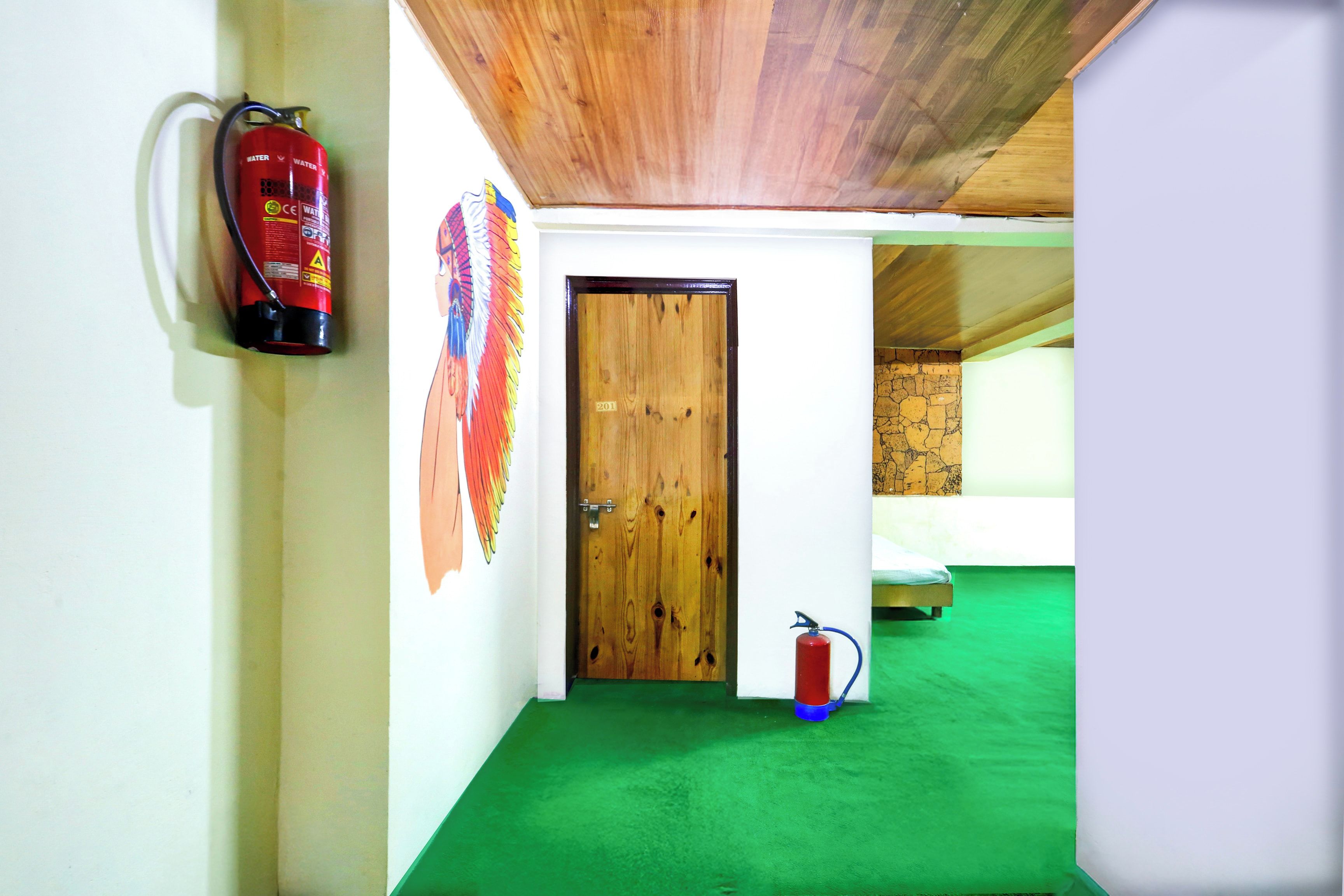 A room with a wooden door, a fire extinguisher mounted on the wall, and a bed with a green carpet.