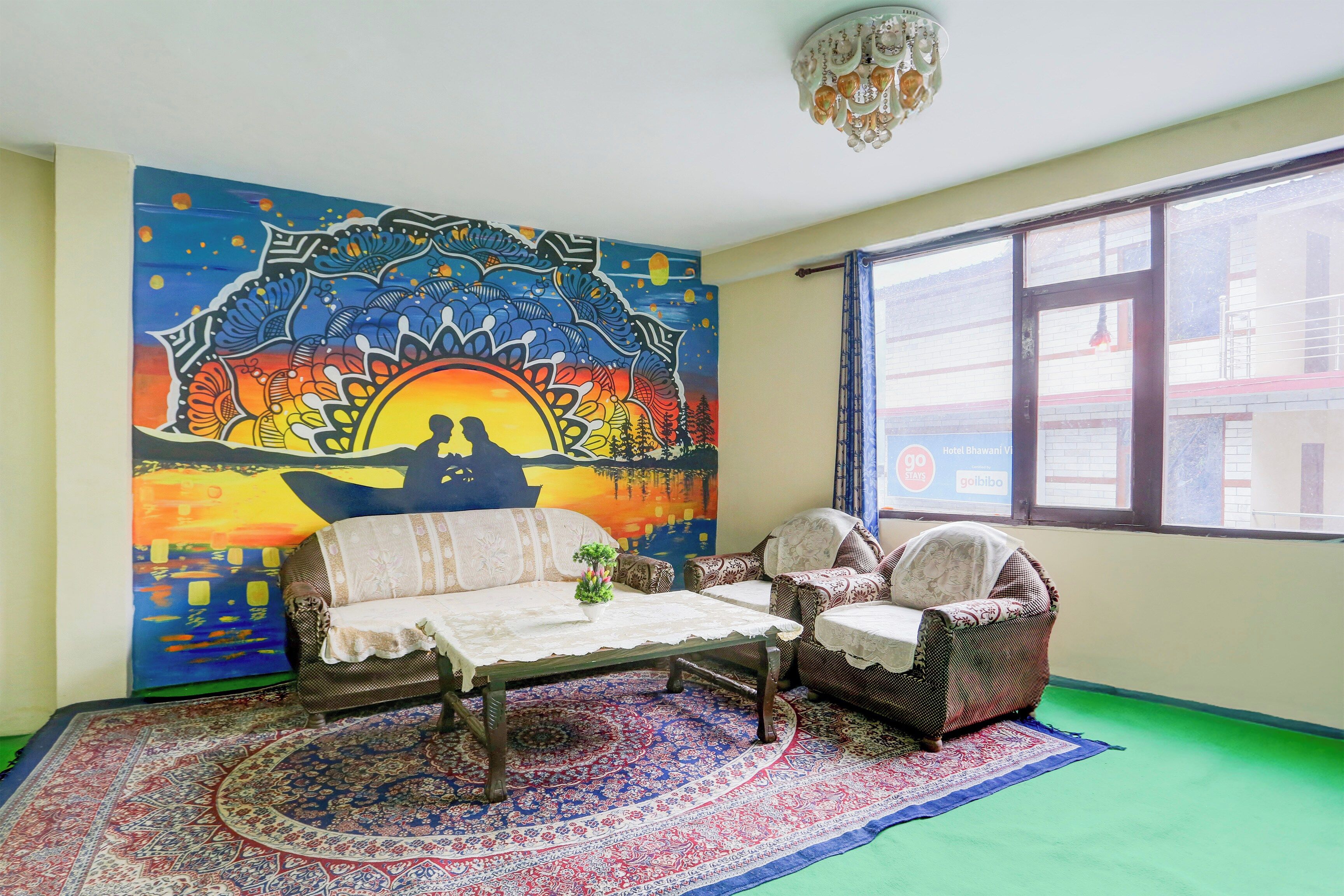 A room with a colorful mural, a sofa, a chair, a small table, and a patterned rug.